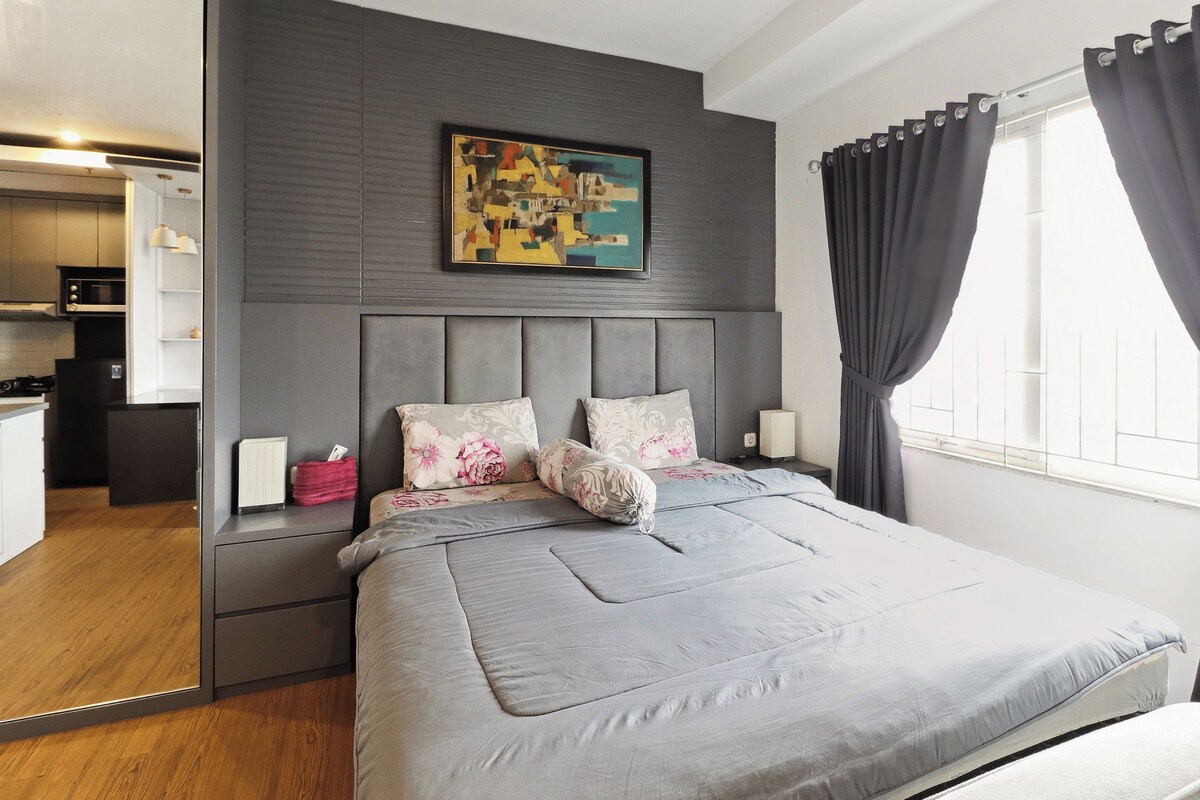 A modern bedroom with a grey headboard, a bed with grey bedding, a painting above the bed, and a window with curtains.