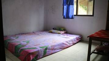 Une petite chambre avec un lit simple, une couette aux couleurs vives, une petite table recouverte d’une nappe rouge et une fenêtre ornée de rideaux bleus.
