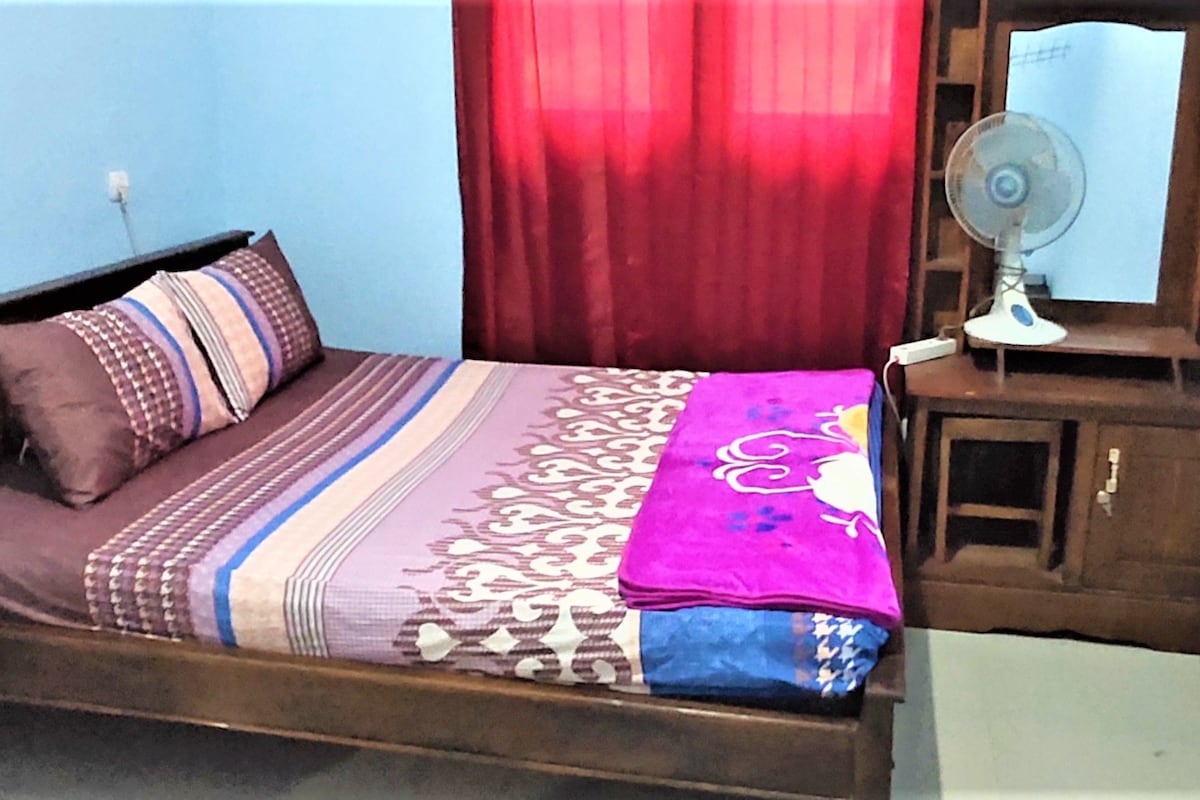 Ein Bett mit gemusterten Betttextilien, ein Nachttisch mit Ventilator und ein Spiegel an der Wand.