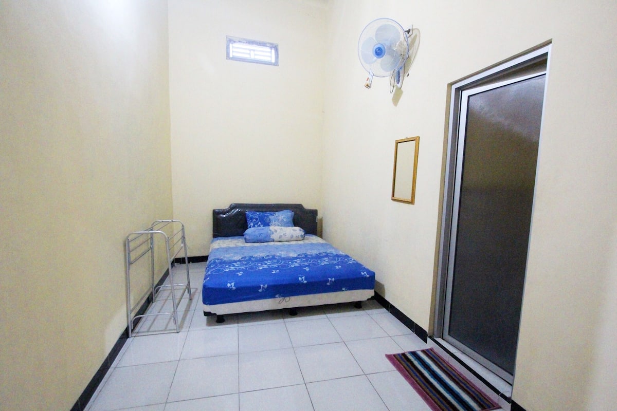A small, narrow room with a bed, a standing fan, a window, and a door.