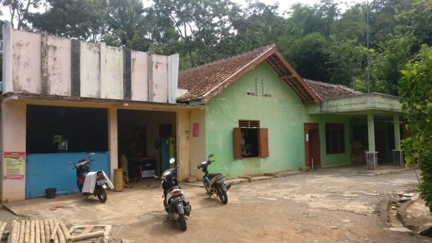 A building with a green facade and a red roof, a blue door, and a motorcycle parked in front. - Adji Homestay - Hostel (Muntuk)