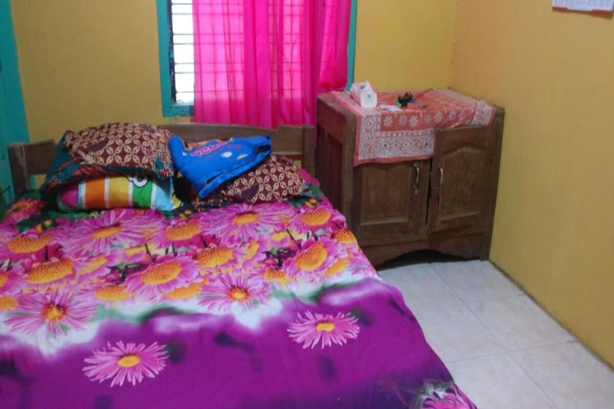 A bedroom with a bed covered in a floral bedspread, a wooden wardrobe, and a window with pink curtains.