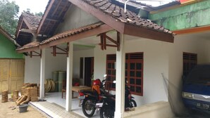 A covered porch with a motorcycle parked in front, a table with items, and a blue car parked to the side. - Wahyu Homestay - Hostel (Muntuk)