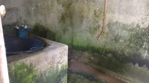 A small, dimly lit room with a concrete sink, a blue container with cleaning supplies, and a single hanging light bulb. - Homestay Miftah Syariah - Hostel (Bantul)