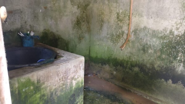 A small, dimly lit room with a concrete sink, a blue container with cleaning supplies, and a single hanging light bulb.