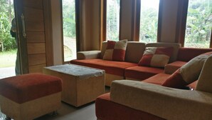 A room with a sofa set and ottoman, large windows with curtains, and a view of greenery outside. - Elvira Homestay - Hostel (Muntuk)