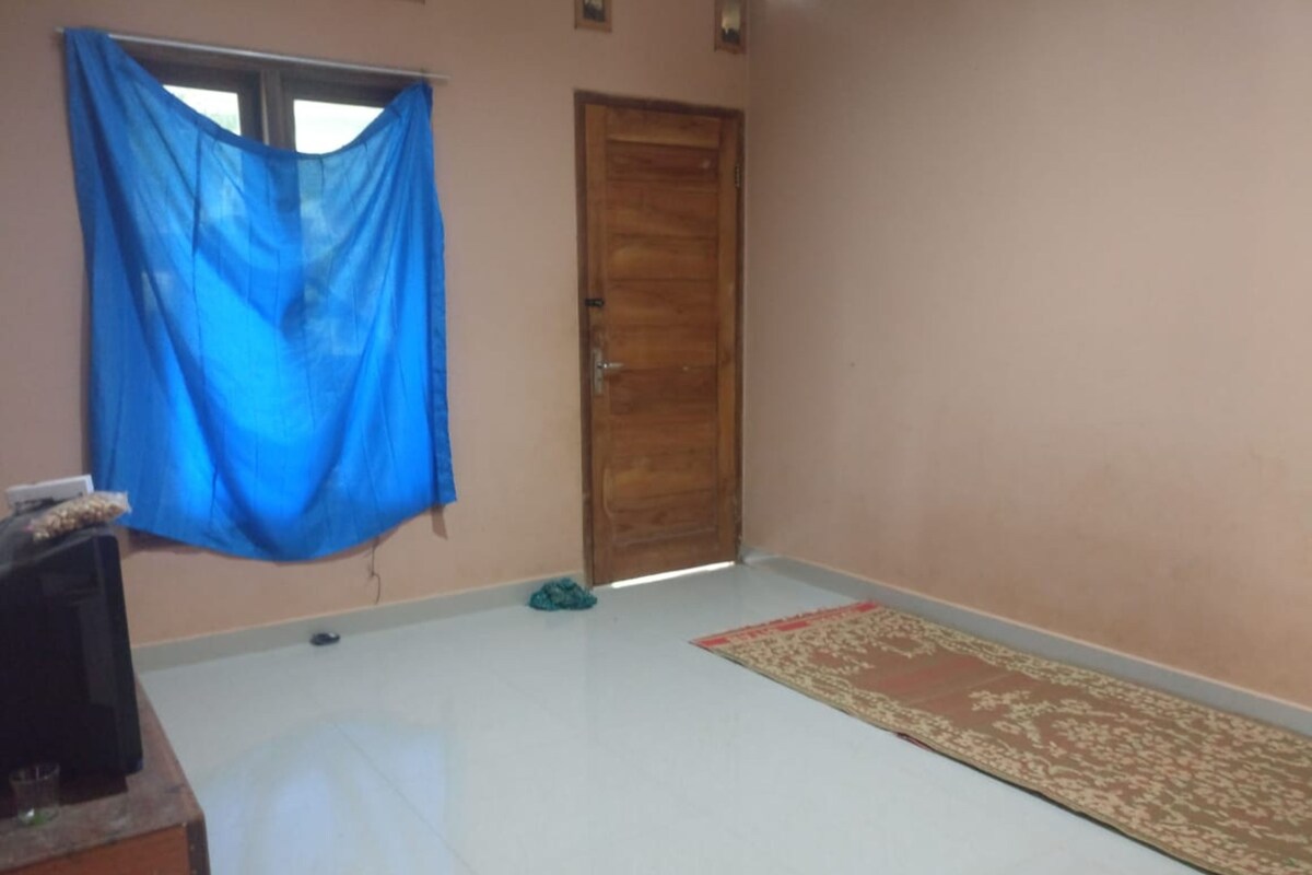 Une pièce avec une porte en bois, un rideau bleu, une petite table avec un verre, et un tapis à motifs posé sur un sol carrelé.