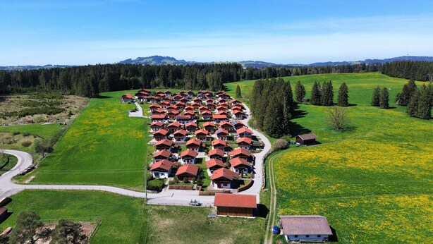 A residential area with houses arranged in rows along a curved road, surrounded by green fields and a forested area.