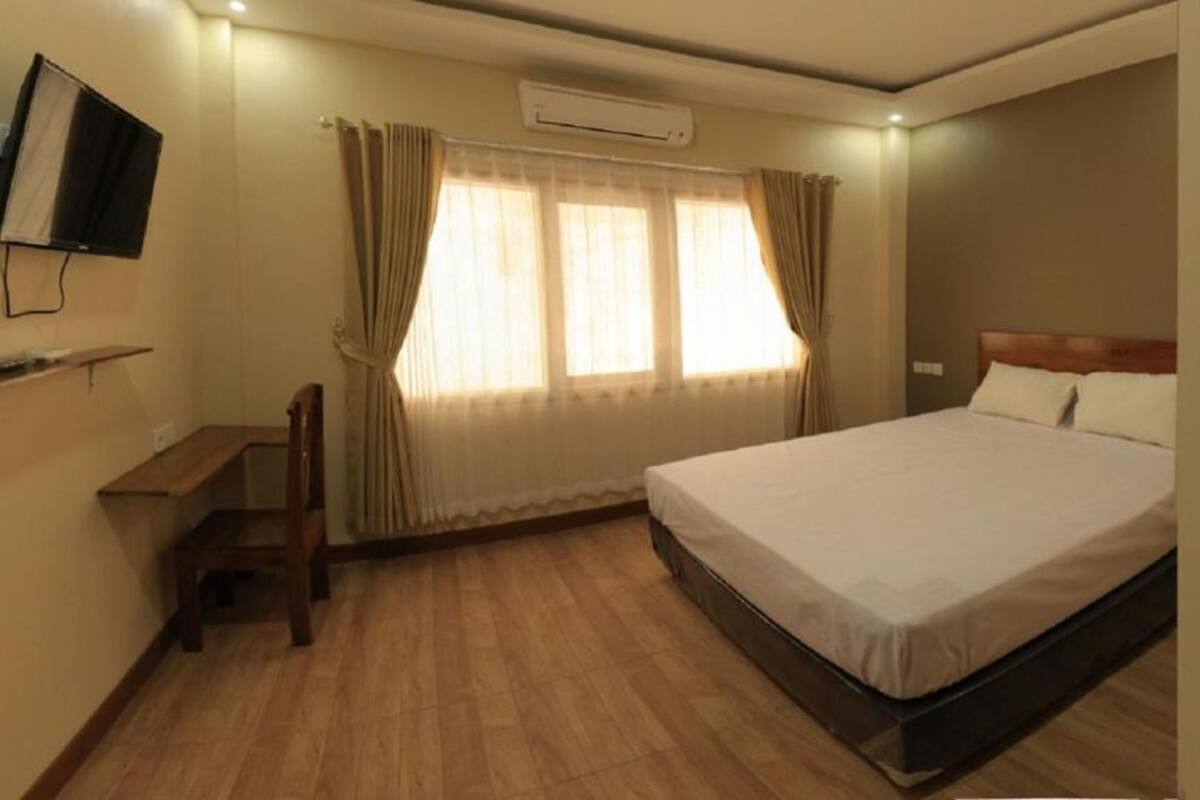 Una camera d'albergo con un letto, una scrivania con una sedia, una televisione a muro e una finestra con tende.