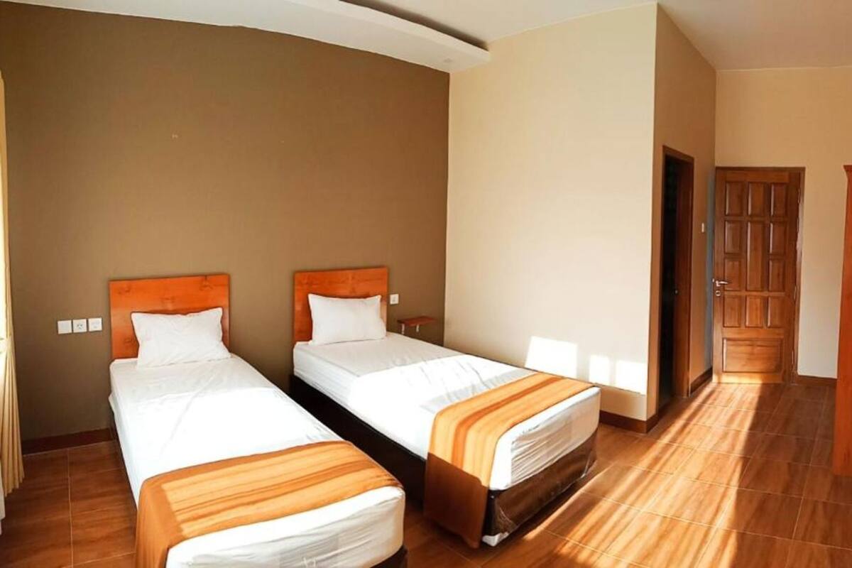 Camera d'albergo con due letti singoli, poggiateste in legno, un'armadio in legno, un televisore a parete e pavimento piastrellato.