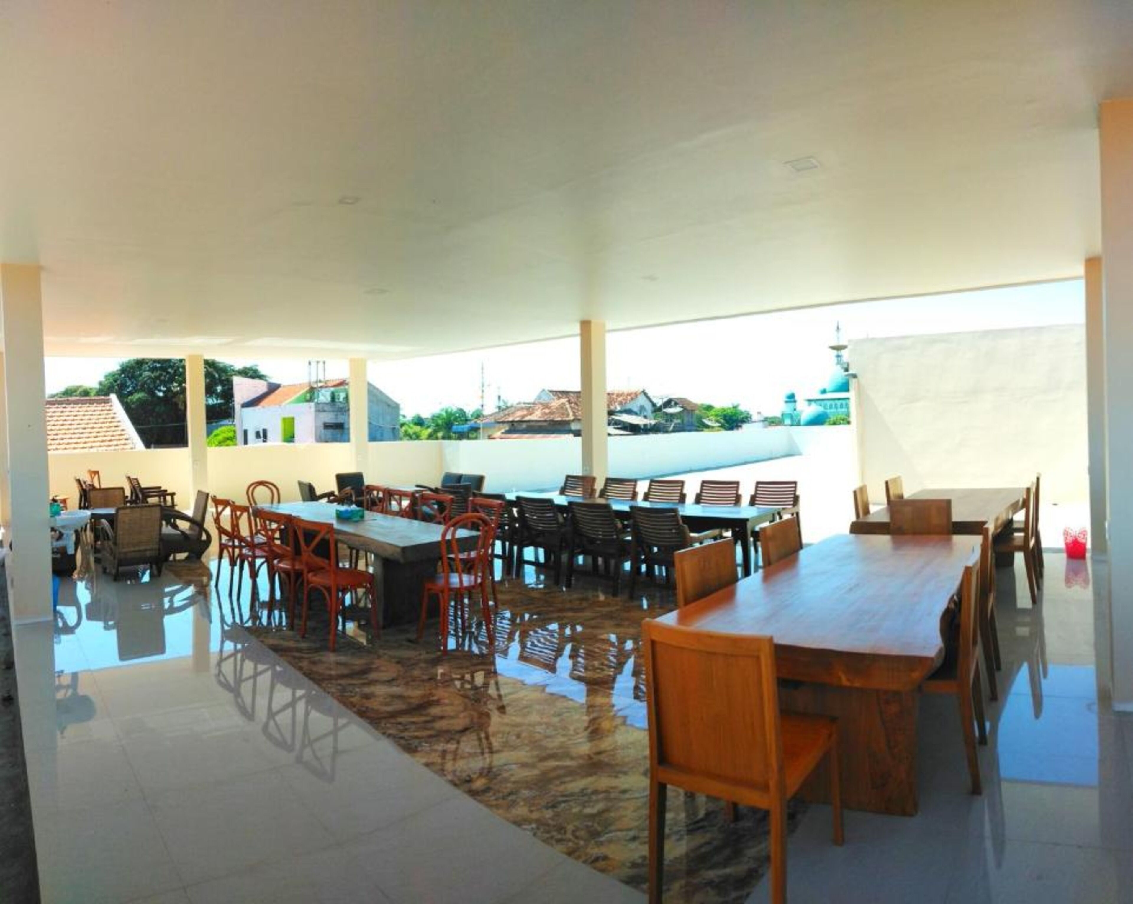 Un vaste espace extérieur aménagé pour les repas, avec de nombreuses tables et chaises en bois, un sol carrelé et une vue sur des bâtiments et une mosquée en arrière-plan.