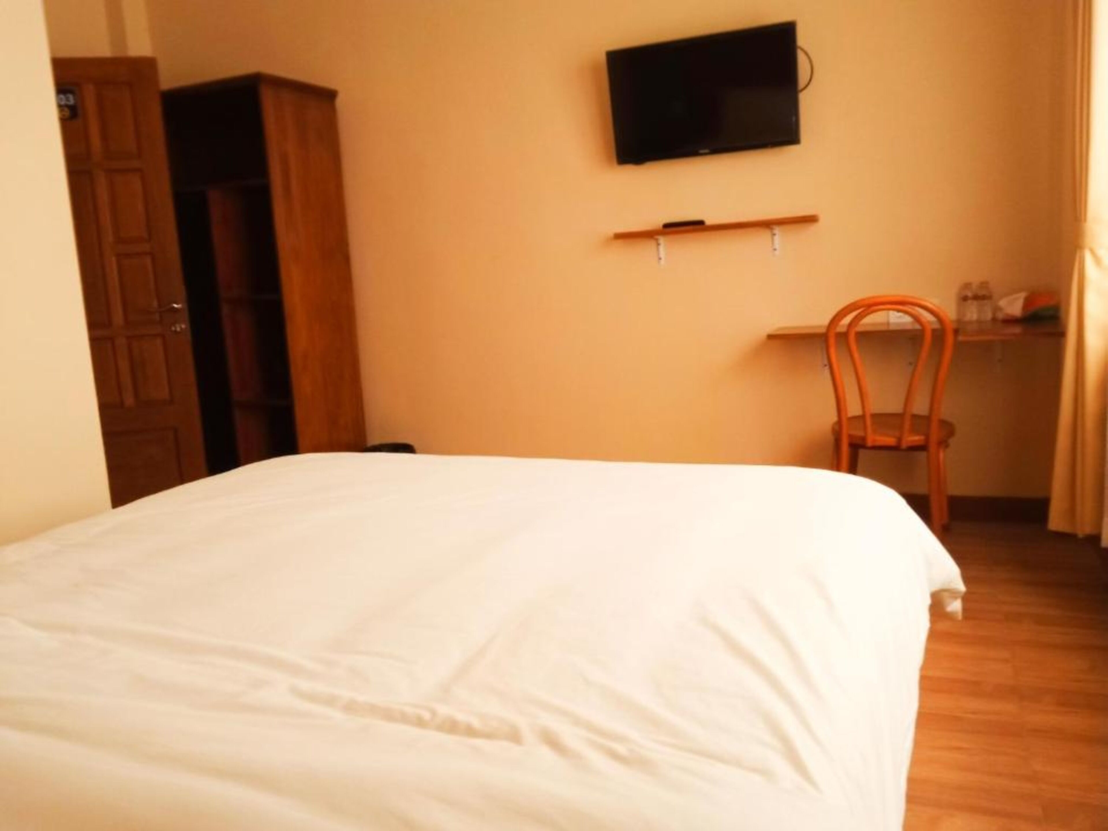 A bedroom with a bed, a TV mounted on the wall, a wooden wardrobe, and a desk with a chair.