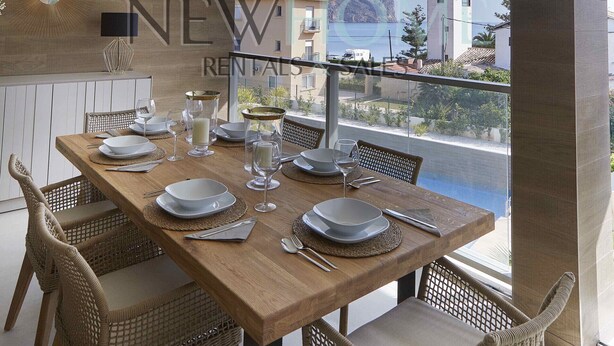 Zona pranzo con tavolo di legno apparecchiato per quattro, sedie in vimini e vista su edifici e sull'acqua.