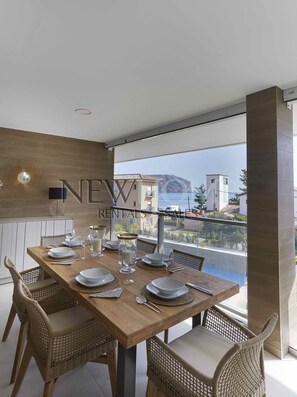 A dining area with a wooden table set for four, wicker chairs, and a view of buildings and water. - Penthouse Duplex Capri (Javea)