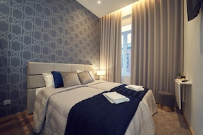 A modern hotel room with a large bed, a bedside table, a window with curtains, and a patterned wall.