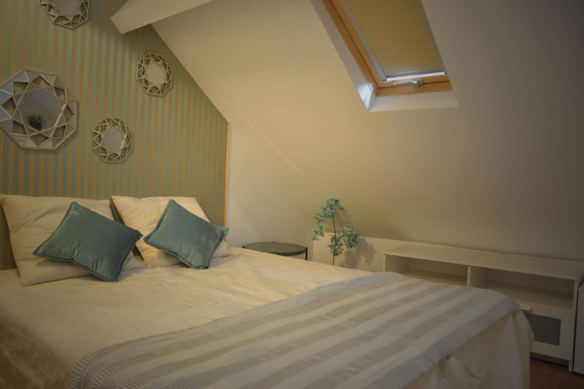 Ein Schlafzimmer mit schrägem Dach, einem Bett mit Kissen, einem Nachttisch und einer kleinen Pflanze.