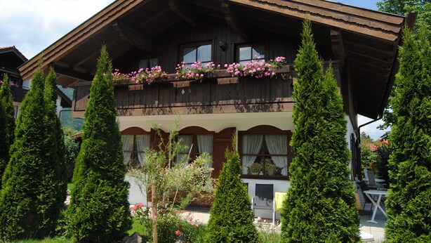 Un tradizionale chalet con un balcone in legno decorato con fiori, circondato da alti alberi sempreverdi.