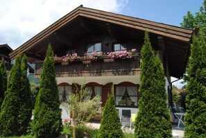 Un chalet traditionnel doté d’un balcon en bois orné de fleurs, entouré de grands arbres à feuilles persistantes.