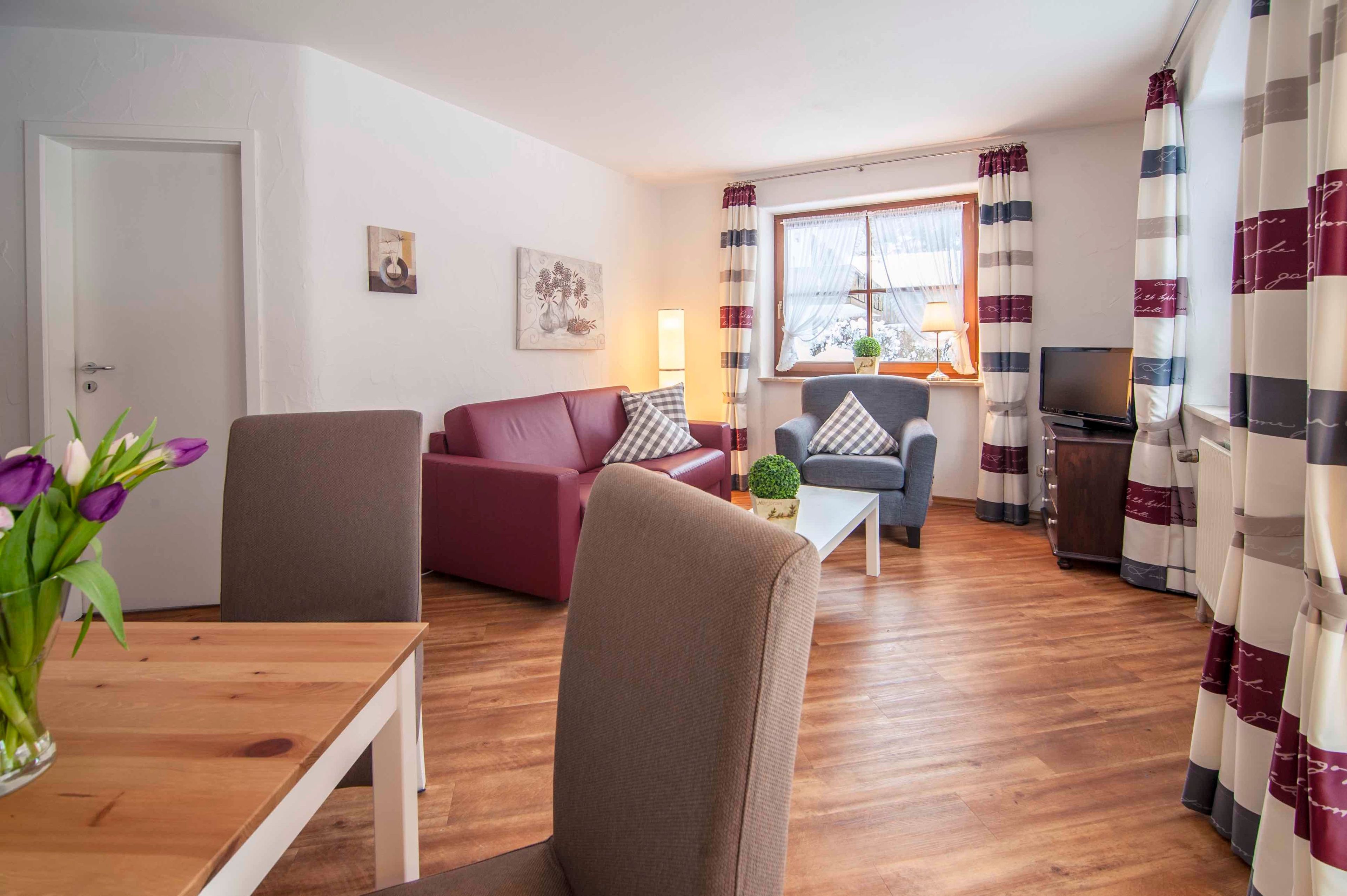 A living room with a sofa, armchairs, a coffee table, and a TV. There is a window with curtains and a door.