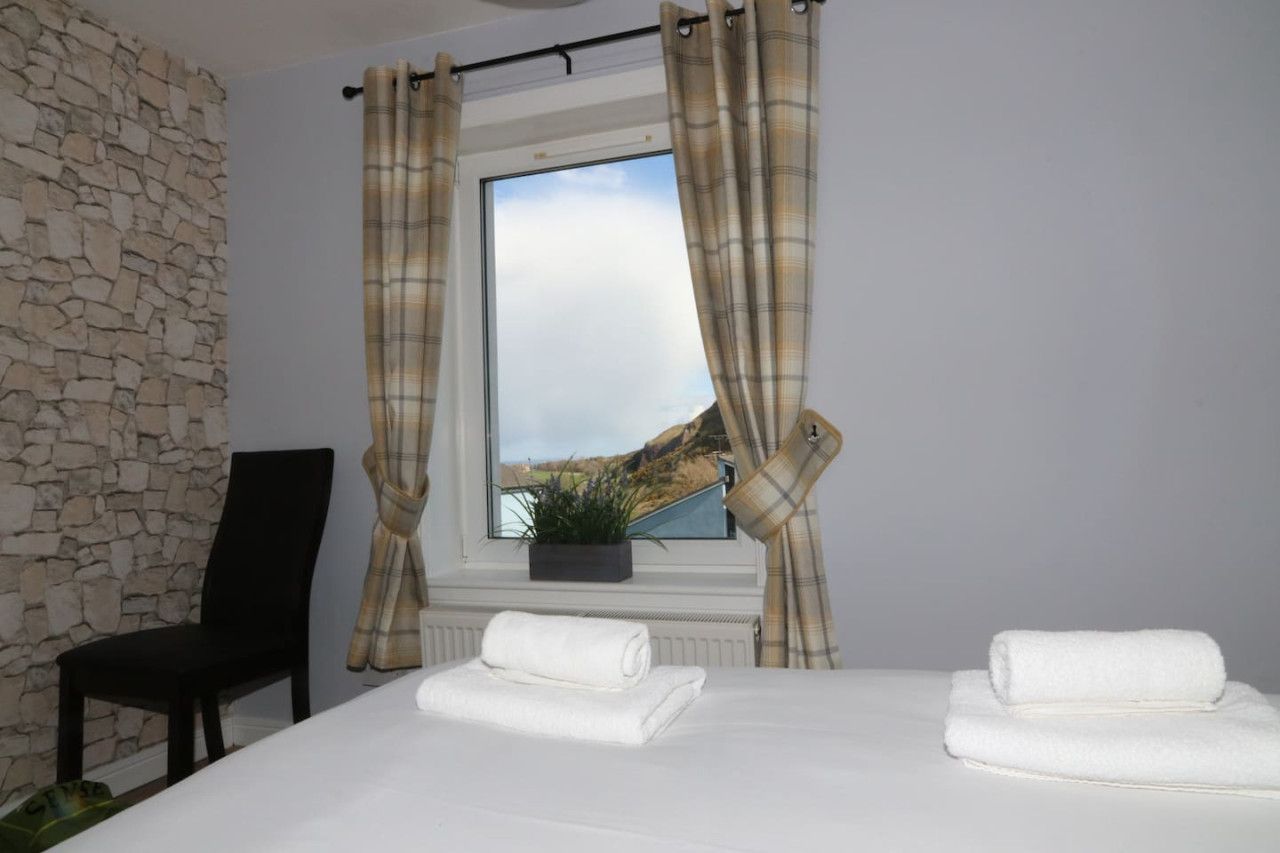 Een hotelkamer met een stenen muur, een bed met witte lakens, twee opgerolde handdoeken, een zwarte stoel en een raam met gordijnen.