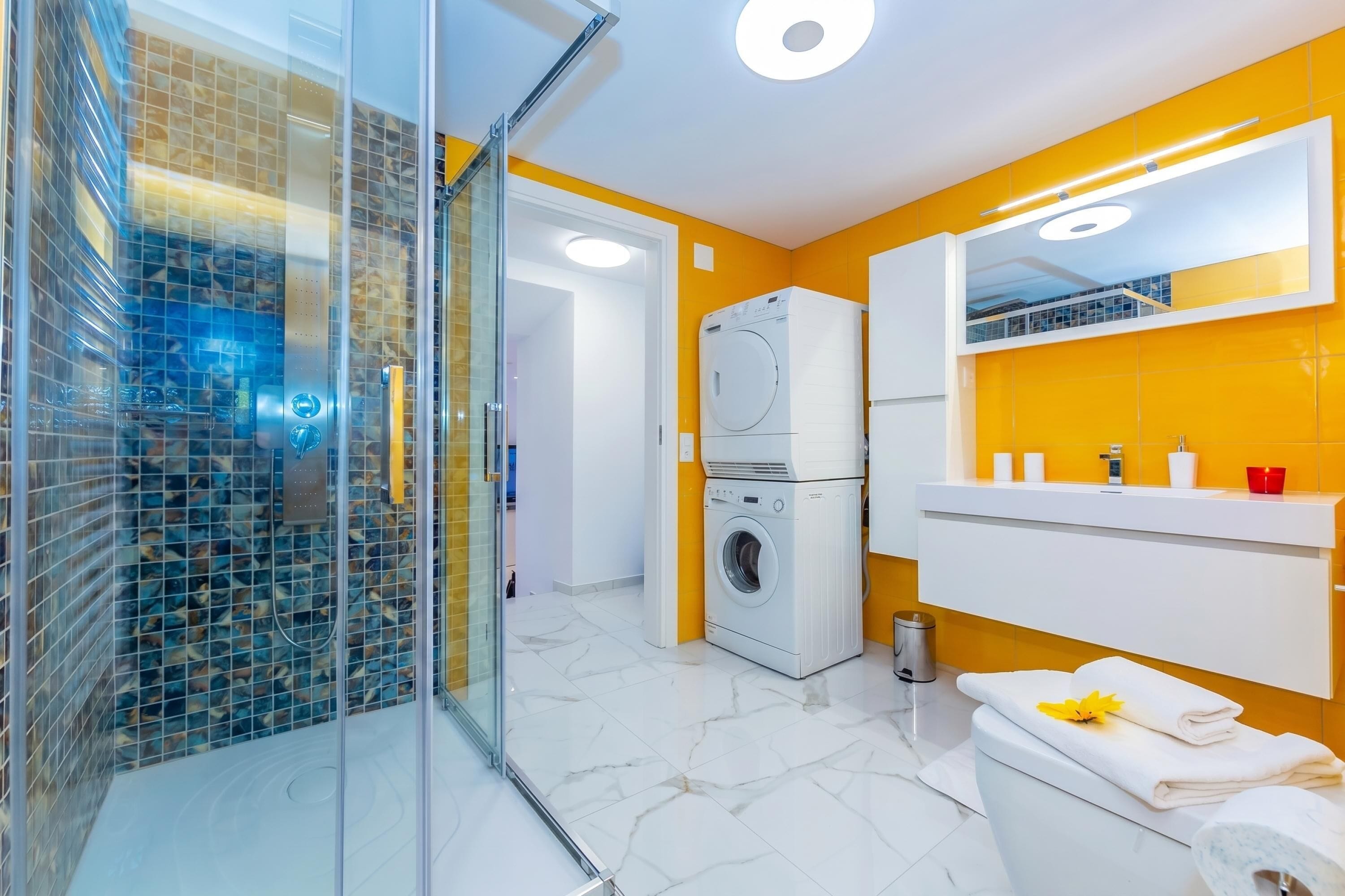 A modern bathroom with a glass shower enclosure, a washing machine, a toilet, and a mirror above the sink.