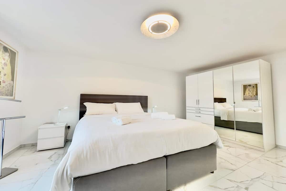 A modern hotel room with a large bed, a bedside table, a nightstand, a wardrobe, and a ceiling light.