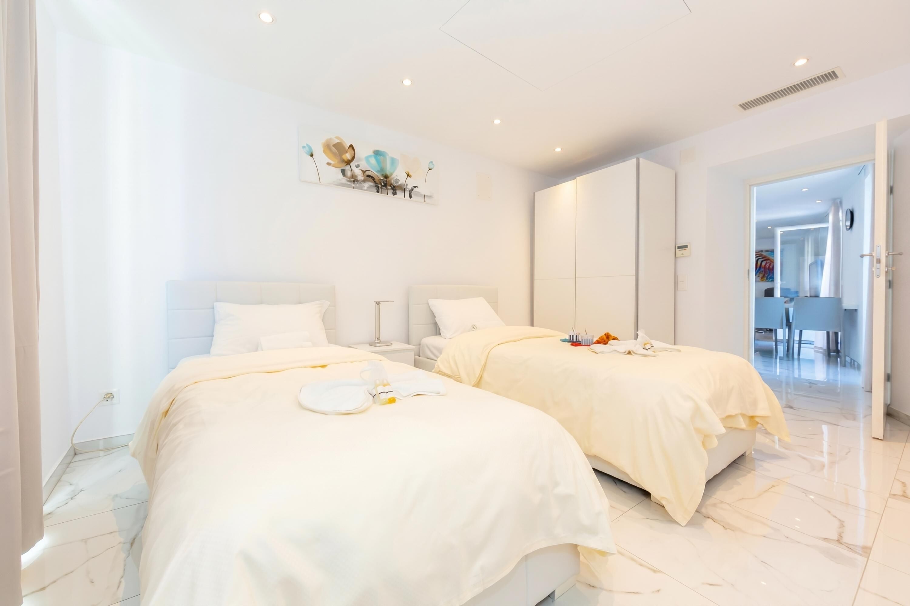 A hotel room with two beds, a marble floor, a wall art piece, and a view into the dining area.
