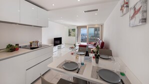 Una cocina moderna con armarios blancos, un comedor con una mesa dispuesta para dos personas y vistas de la ciudad a través de amplios ventanales.