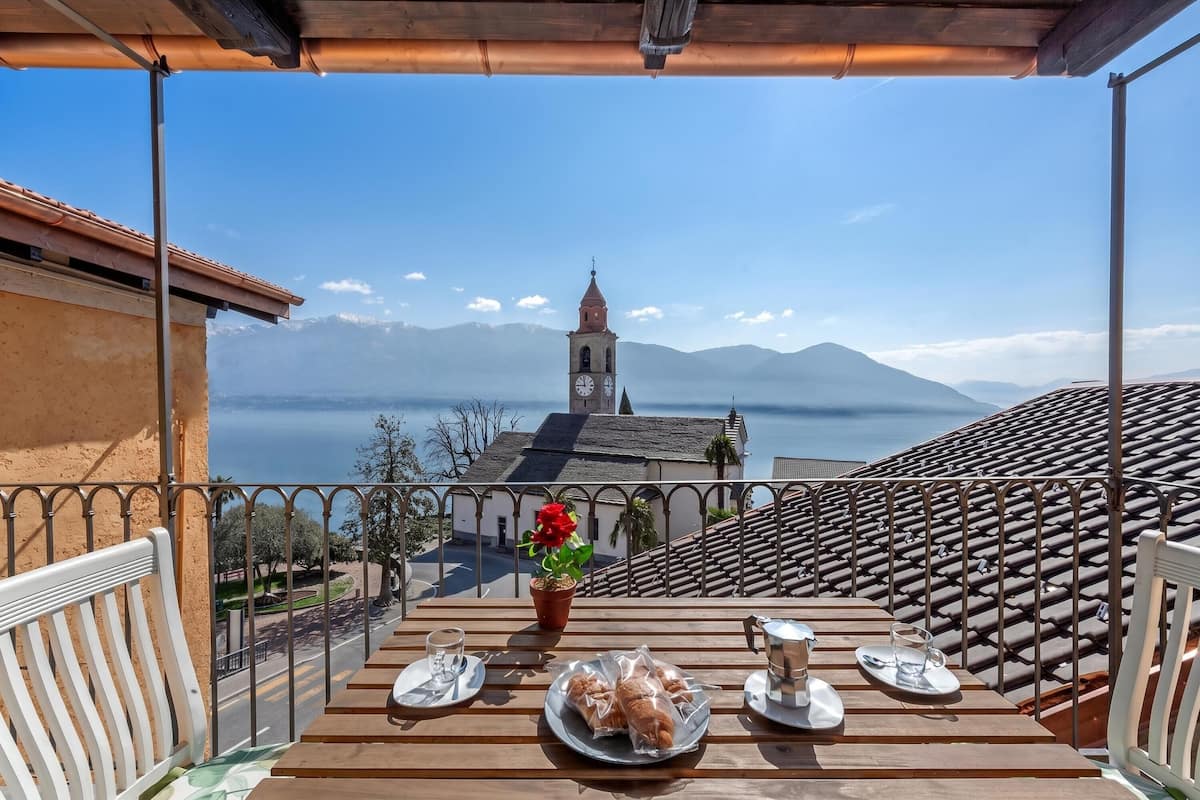 A balcony with a table set for two, overlooking a town with a church and mountains.