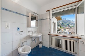 A bathroom with two sinks, a bidet, a mirror, a radiator, and a window with a view of a residential area.