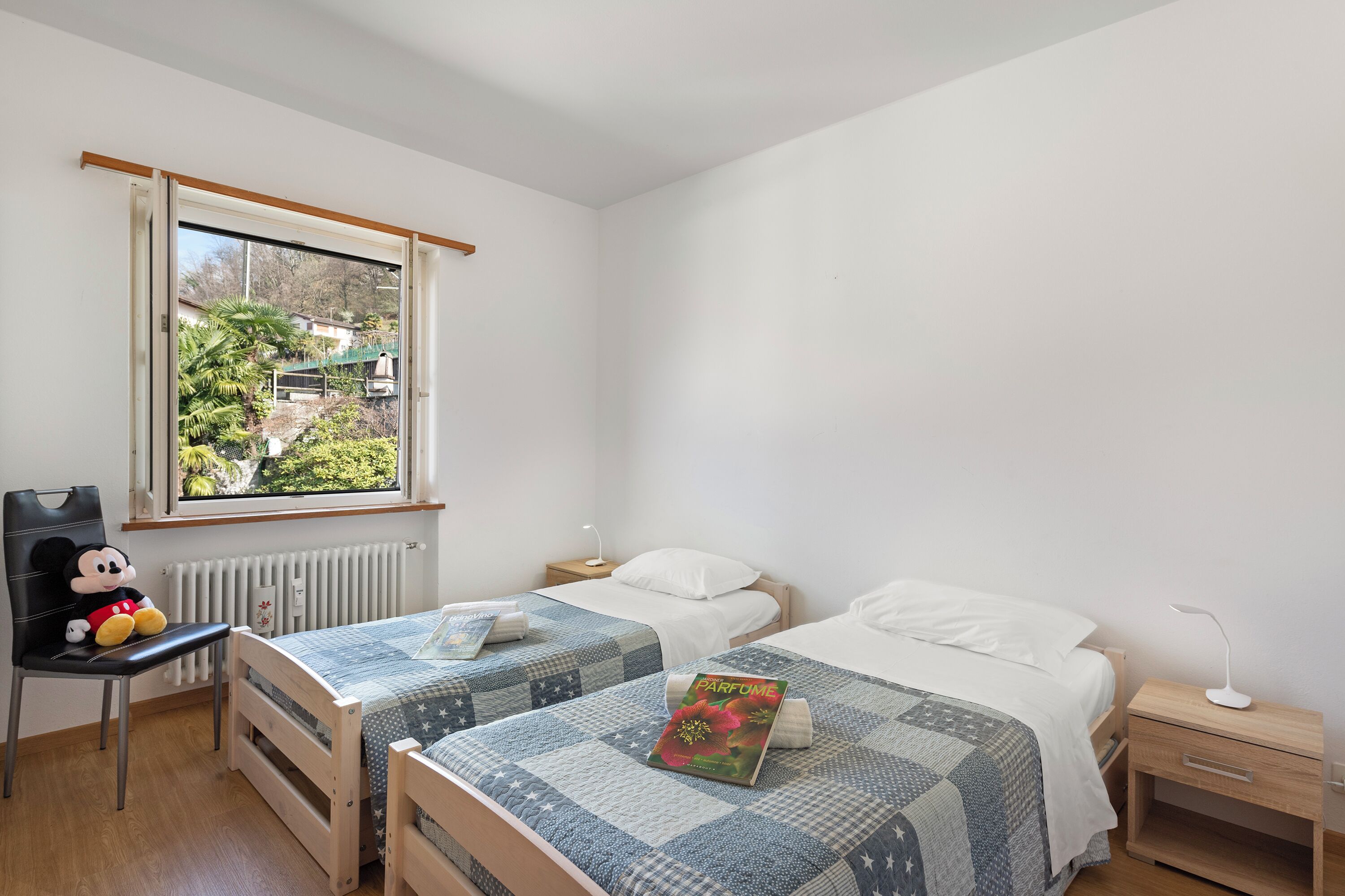 A bedroom with two beds, a Mickey Mouse chair, a radiator, and a window with a view of houses and greenery.