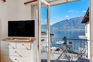 Un balcón con televisor, una mesa con un jarrón de flores y vistas a un lago y montañas.