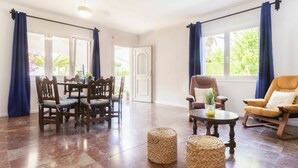 Un salon spacieux avec un coin repas, de grandes fenêtres ornées de rideaux bleus et un ventilateur de plafond.