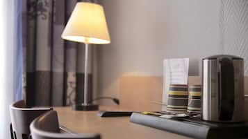 A hotel room with a desk, a lamp, a coffee maker, and a stack of books.
