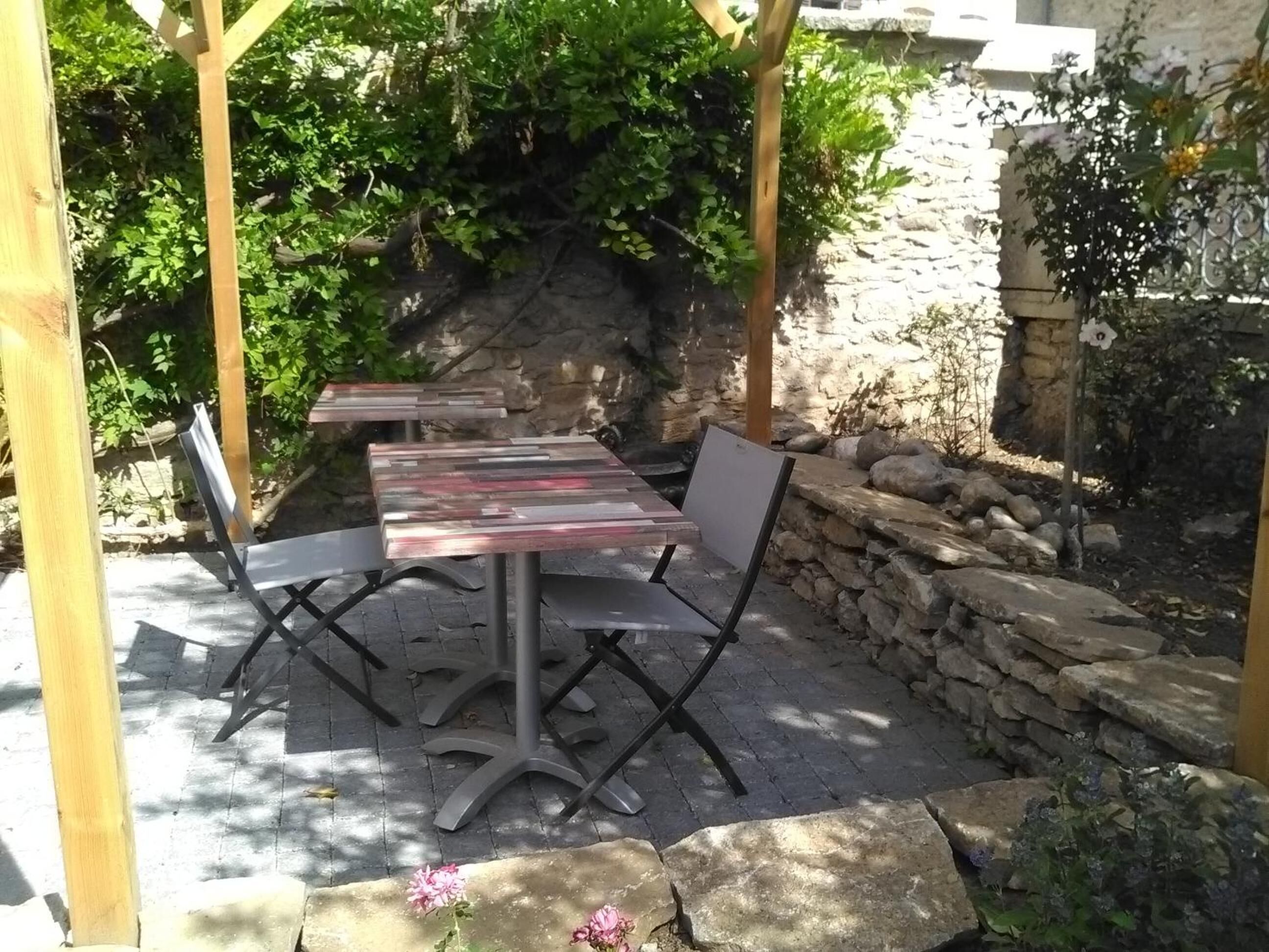Un patio al aire libre con mesas y sillas de madera, rodeado de muros de piedra y follaje.