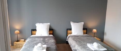 Habitación doble con dos camas, cada una con cabecero de madera y colcha gris. Hay almohadas blancas y toallas enrolladas en cada cama. Un candelabro cuelga sobre las camas.