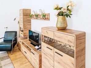 A modern living room with a wooden TV stand, a flat-screen TV, a glass cabinet with wine glasses, a blue chair, and a vase with flowers on a wooden cabinet.