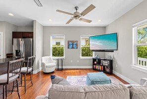 A modern living room with a flat-screen TV, a ceiling fan, and a dining area with a refrigerator and bar stools.