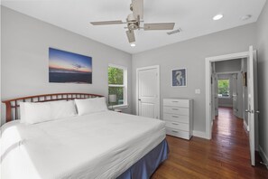 A bedroom with a bed, a ceiling fan, a window with a view, a door, and a hallway.