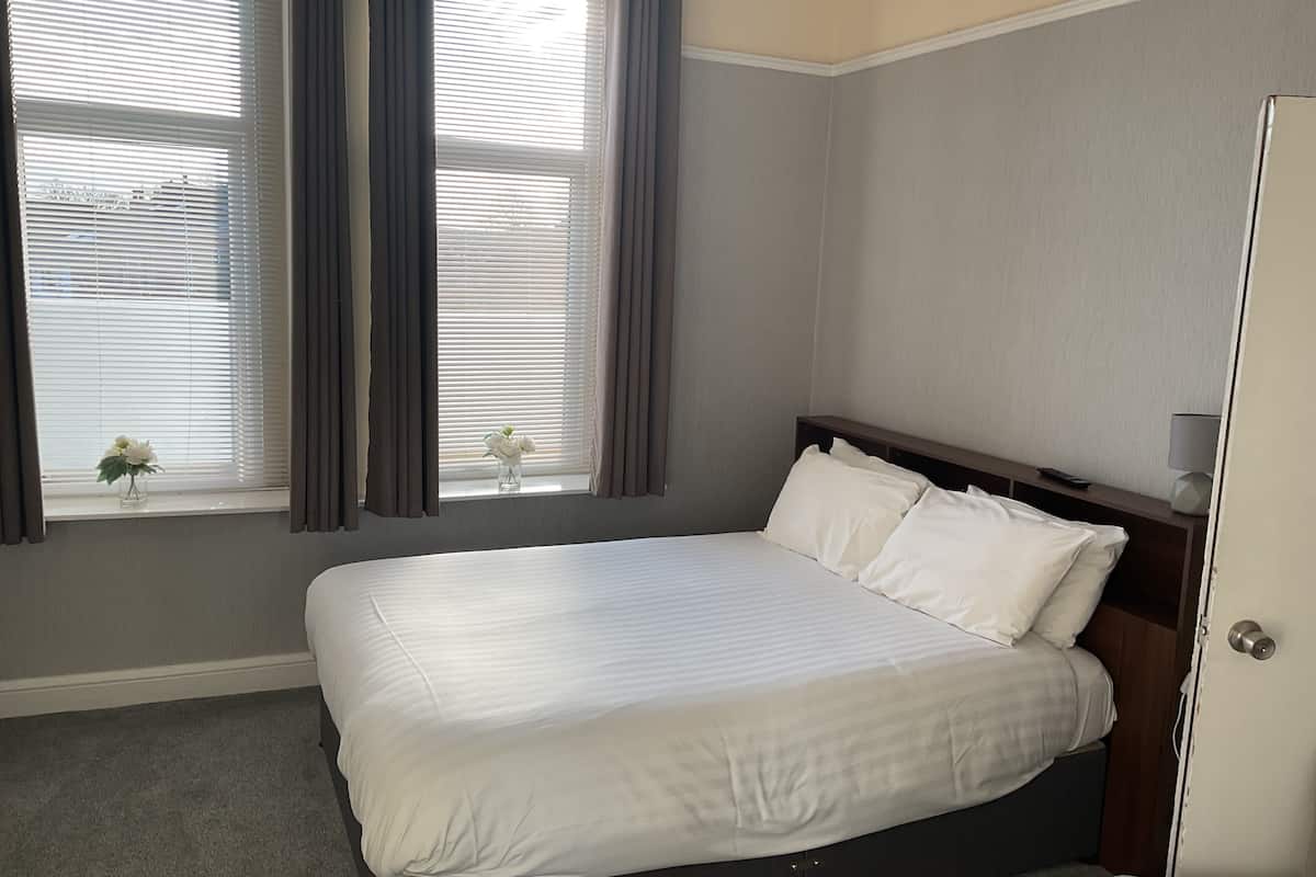 A neatly made bed with white linens in a room with grey walls and a window with curtains.