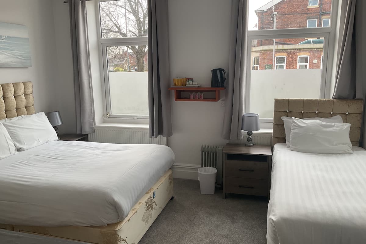 A bedroom with two beds, a radiator, a bedside table with a lamp, a shelf with items, and a window with curtains.