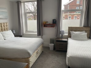 A bedroom with two beds, a radiator, a bedside table with a lamp, a shelf with items, and a window with curtains. - Claremont Hotel (Preston)
