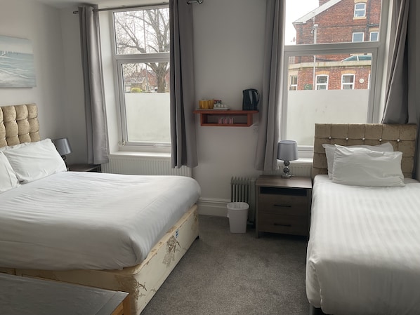 Un dormitorio con dos camas, un radiador, una mesita de noche con una lámpara, una estantería con objetos y una ventana con cortinas.