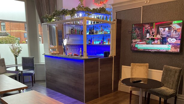 A bar area with a counter, shelves of bottles, and a television screen displaying a show. There are chairs and tables around the bar. - Claremont Hotel (Preston)