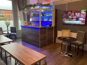 Zona de bar con mostrador, estantes llenos de botellas y una pantalla de televisión mostrando un programa. Hay sillas y mesas alrededor de la barra.