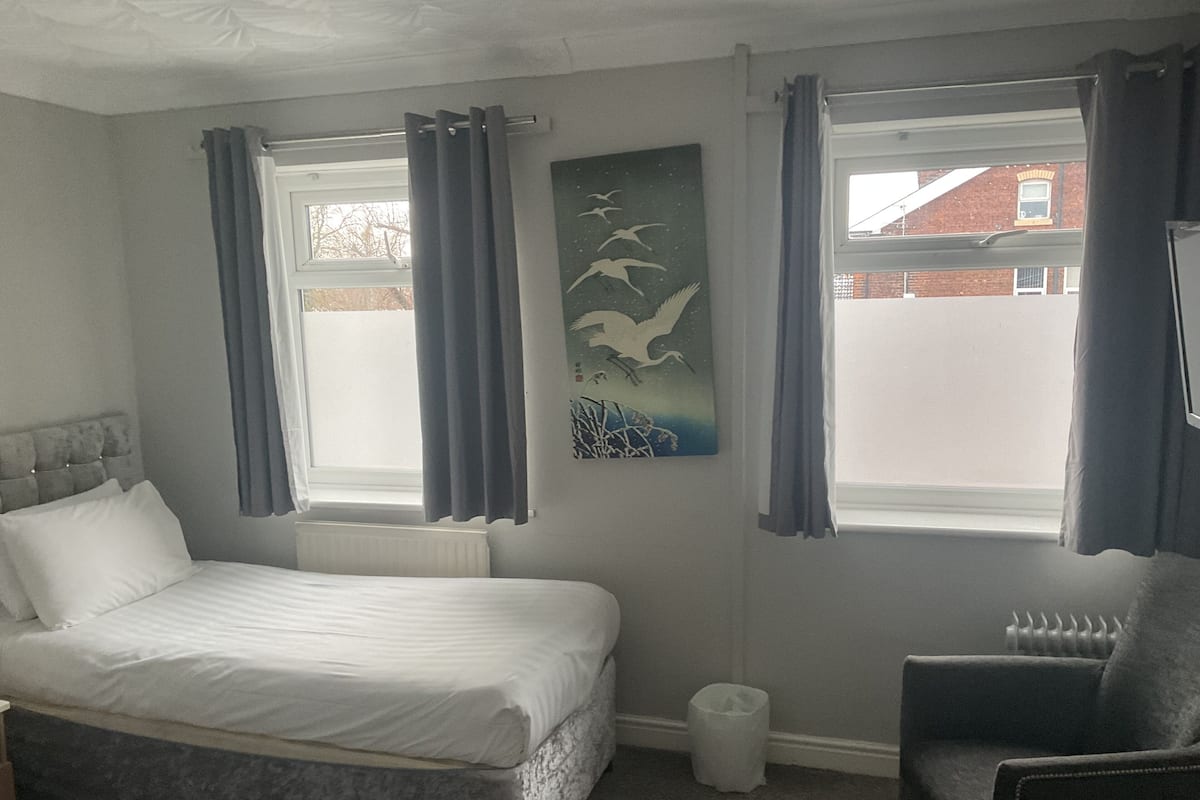 A bedroom with a bed, a chair, a radiator, a window with curtains, and a painting on the wall.