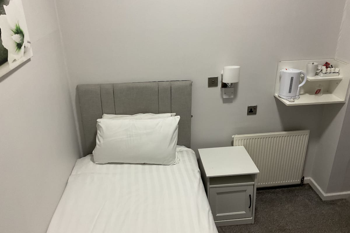 A small, neatly made hotel room with a single bed, a bedside table, a radiator, and a shelf with a kettle and cups.