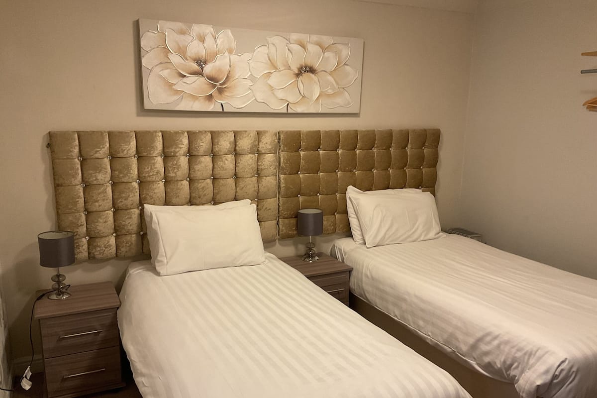 A hotel room with two single beds, a large tufted headboard, a nightstand with a lamp, and a floral artwork on the wall.