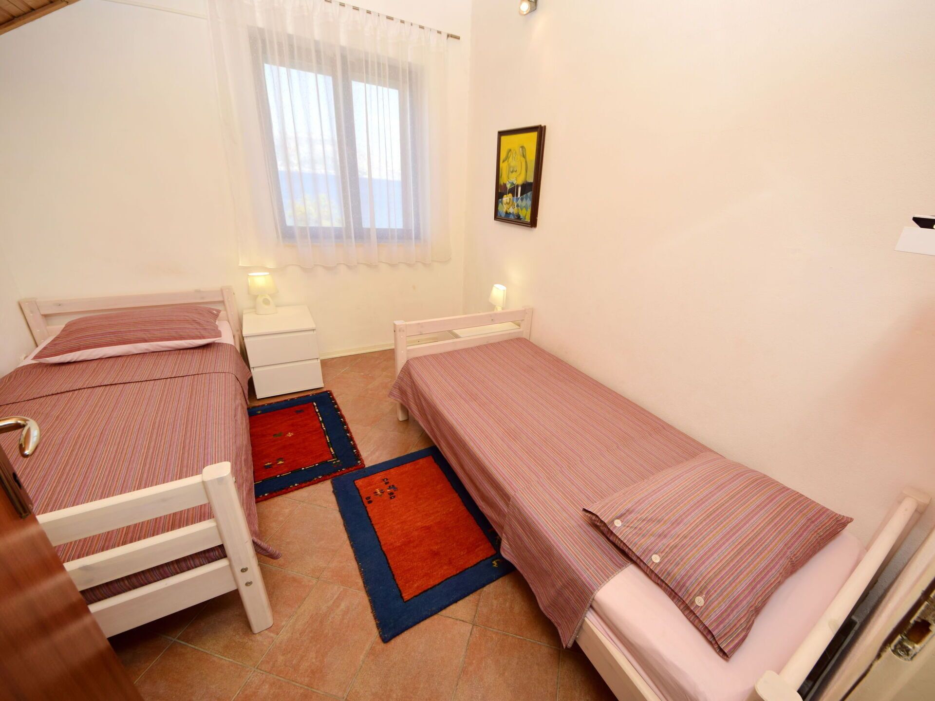 A small, clean room with two single beds, a nightstand, a red and blue rug, and a window with sheer curtains.