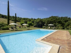 Ein rechteckiger Swimmingpool mit einer Ziegelterrasse darum. Liegestühle und Sonnenschirme befinden sich rund um den Pool.